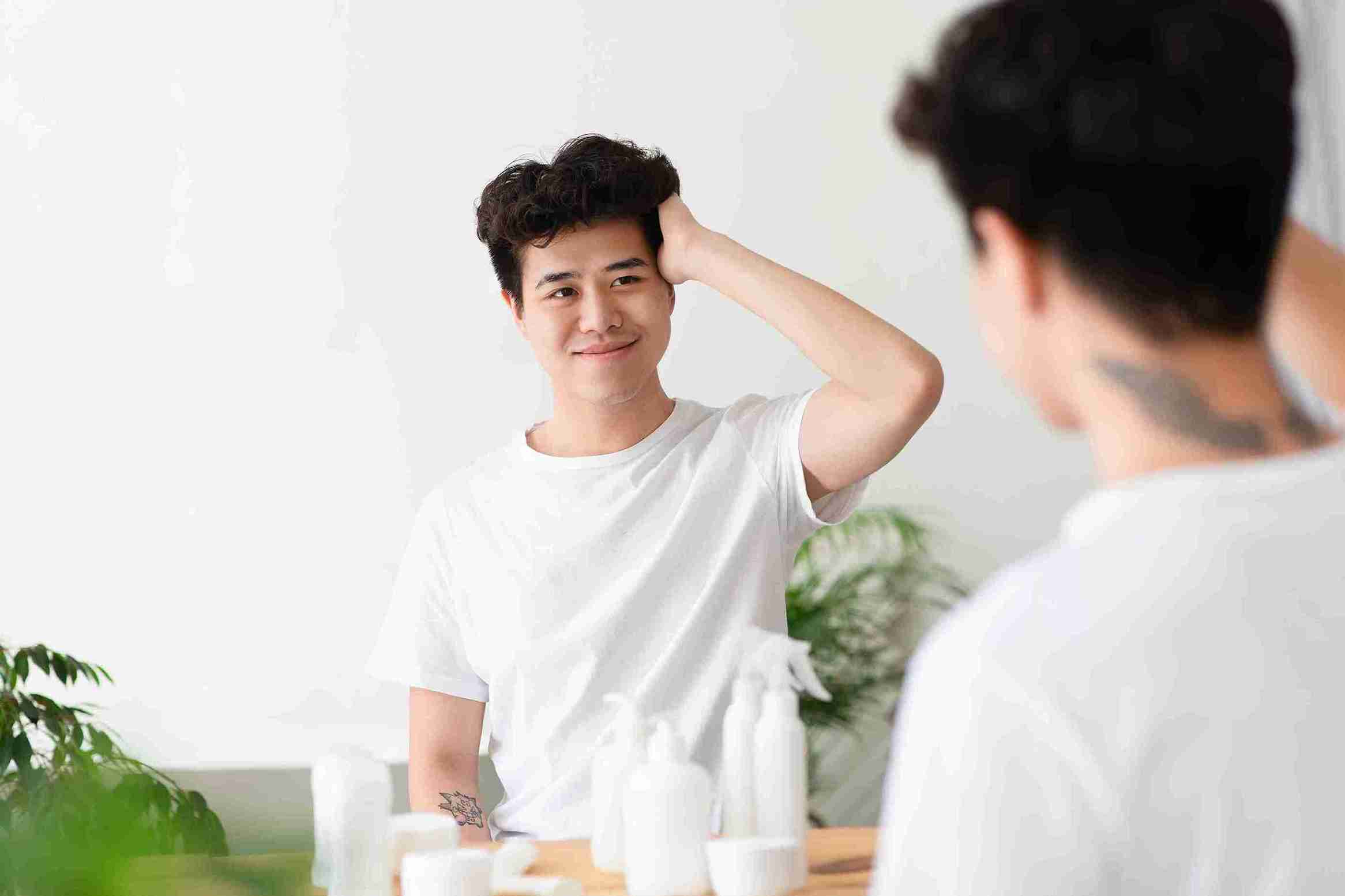 A young man smiling while looking at his reflection in the mirror, touching his hair, surrounded by hair care products—demonstrating confidence in hair care routines. Thinking about how to Strengthen Hair.