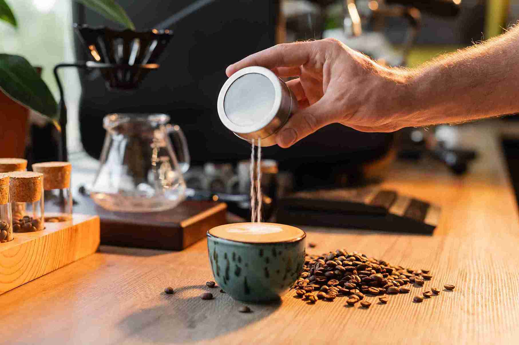 A hand pours sugar into a cup of coffee next to scattered beans, symbolizing the caution or restriction of drinking coffee after hair transplant.