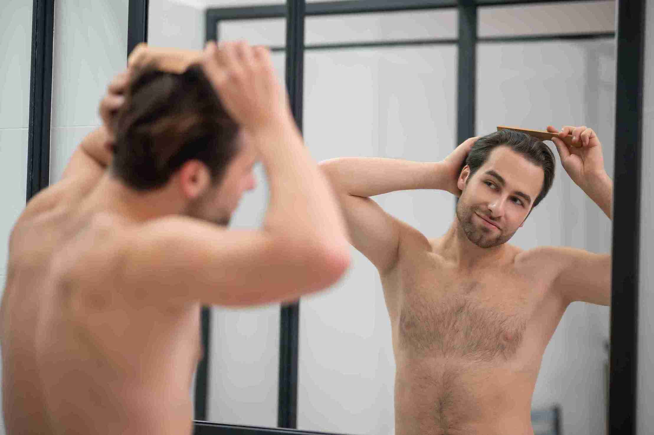A young man standing shirtless in front of a bathroom mirror, brushing his hair and checking his appearance, representing grooming and the question 'Is a Hair Transplant Permanent?