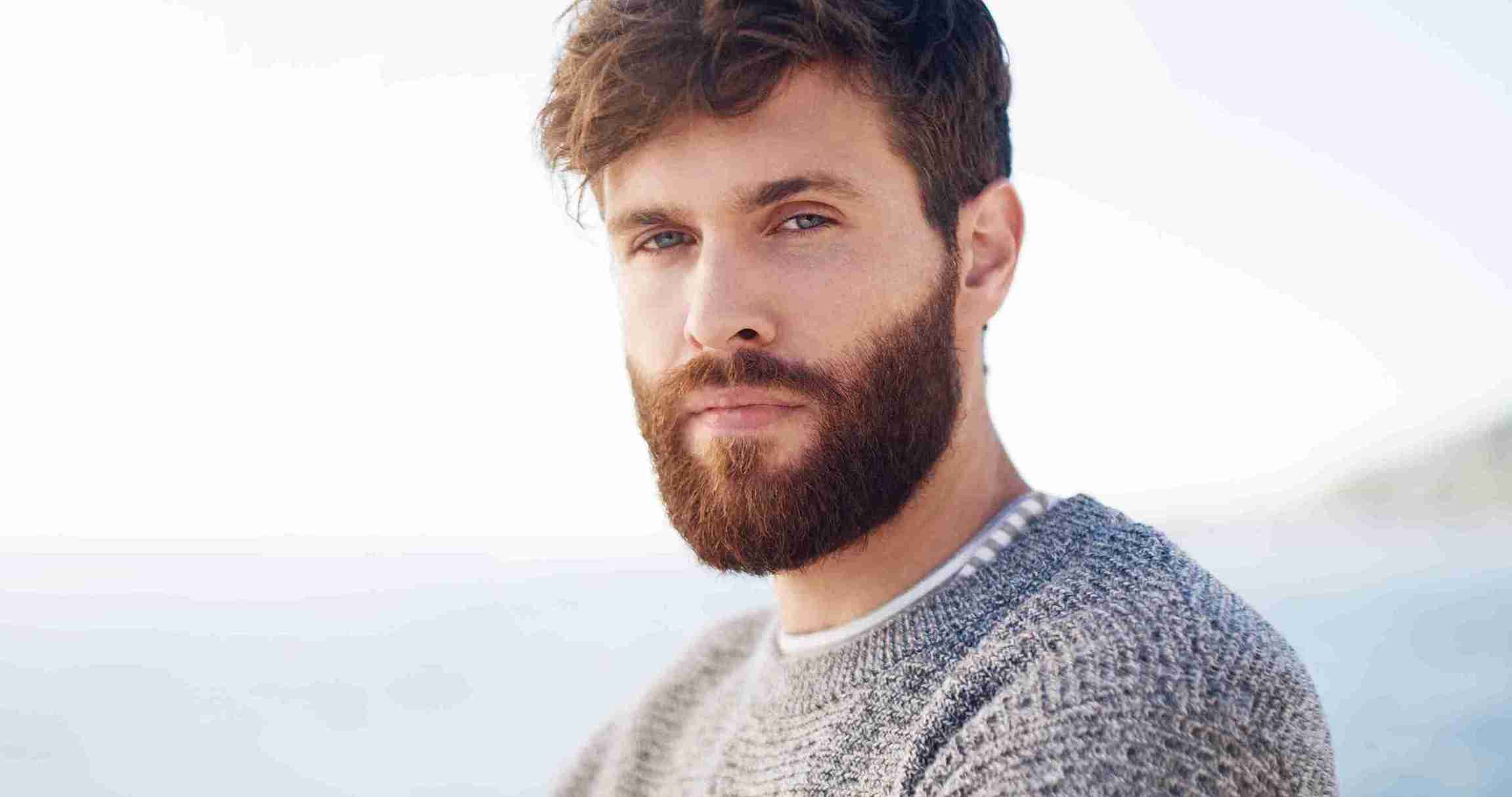 Porträt eines Mannes mit vollem Bart und kurzen Haaren am Meer – Symbolbild für das Thema Lücken im Bart und Bartpflege.