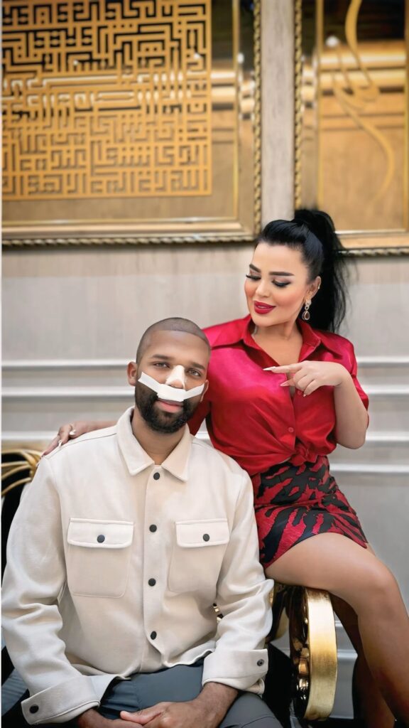 A man with a nasal bandage is sitting on a chair, while Asli Tarcan is sitting next to him, smiling and pointing at him.