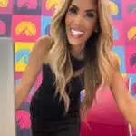 Sandra Ali smiling and leaning forward over a desk, wearing a sleeveless black dress in a brightly colored studio setting, used in an article discussing Sandra Ali’s weight loss surgery.