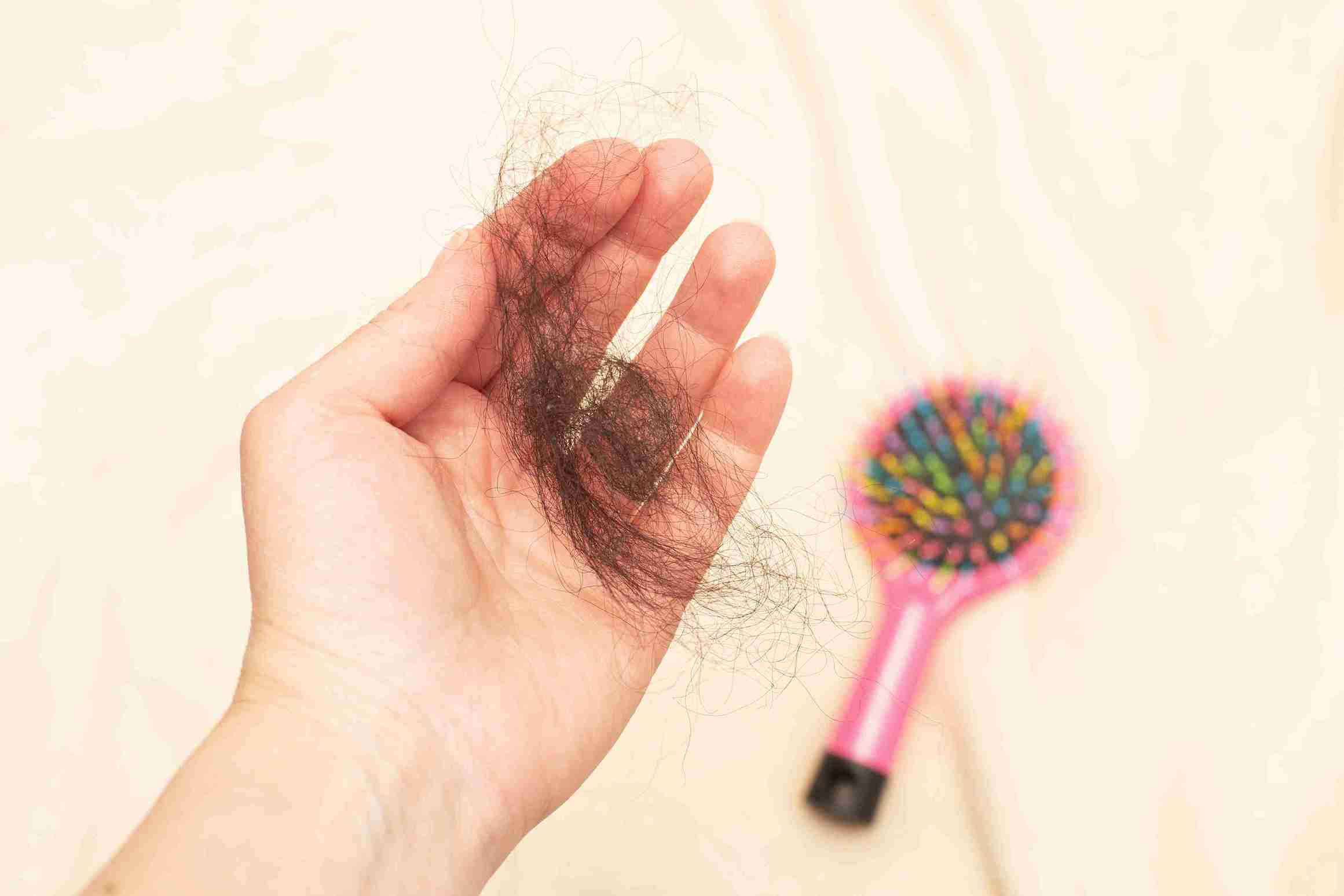 A hand holding a clump of shed hair, with a colorful hairbrush blurred in the background, illustrating Hormonal hair loss and excessive hair shedding.