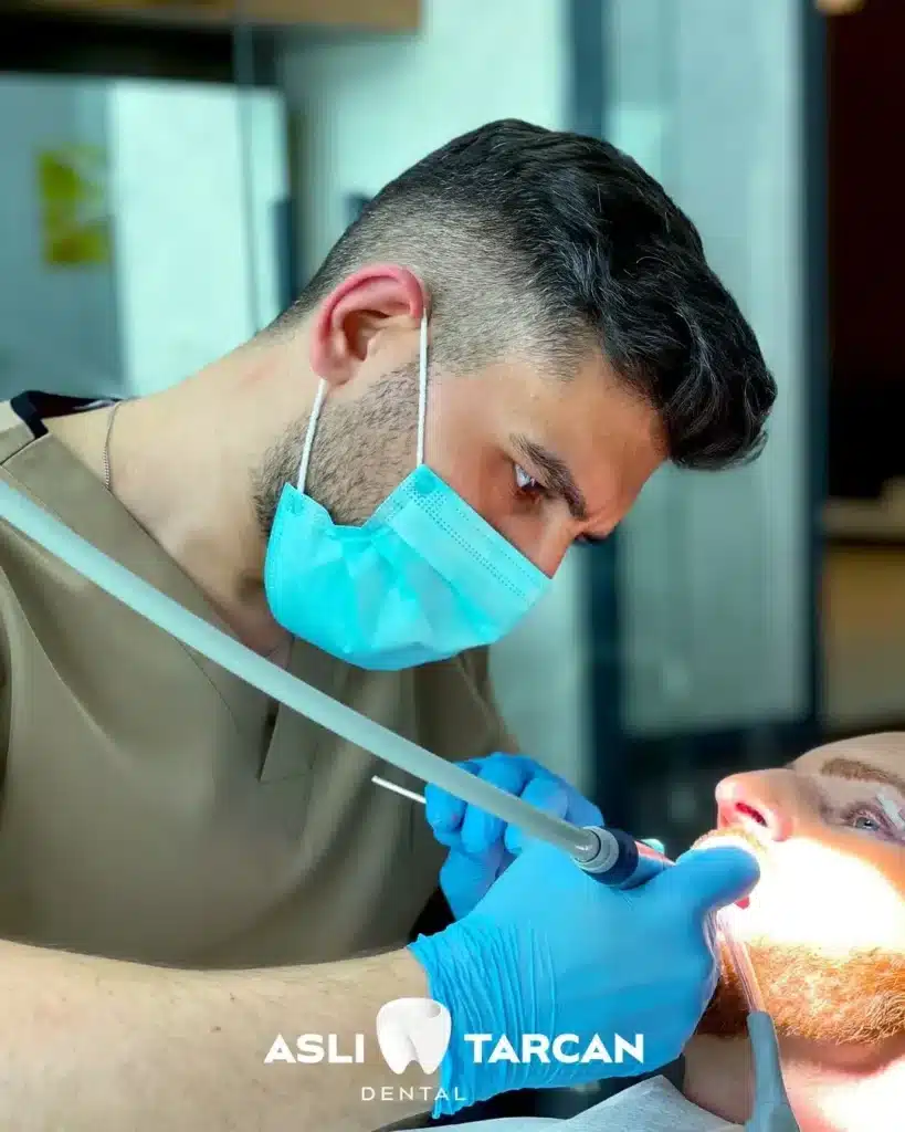 Zahnarzt mit Mundschutz und blauen Handschuhen behandelt einen liegenden Patienten; Nahaufnahme einer Zahnbehandlung mit zahnärztlichen Instrumenten in einer modernen Praxis, unten mit Logo einer Zahnklinik.