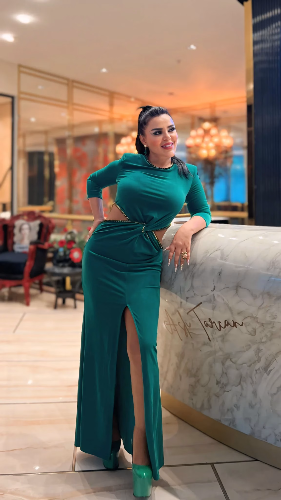 Mujer elegante con vestido verde esmeralda posando en un interior lujoso junto a un mostrador de mármol, transmitiendo confianza y estilo, asociada a servicios de Trasplante Capilar Turquía.