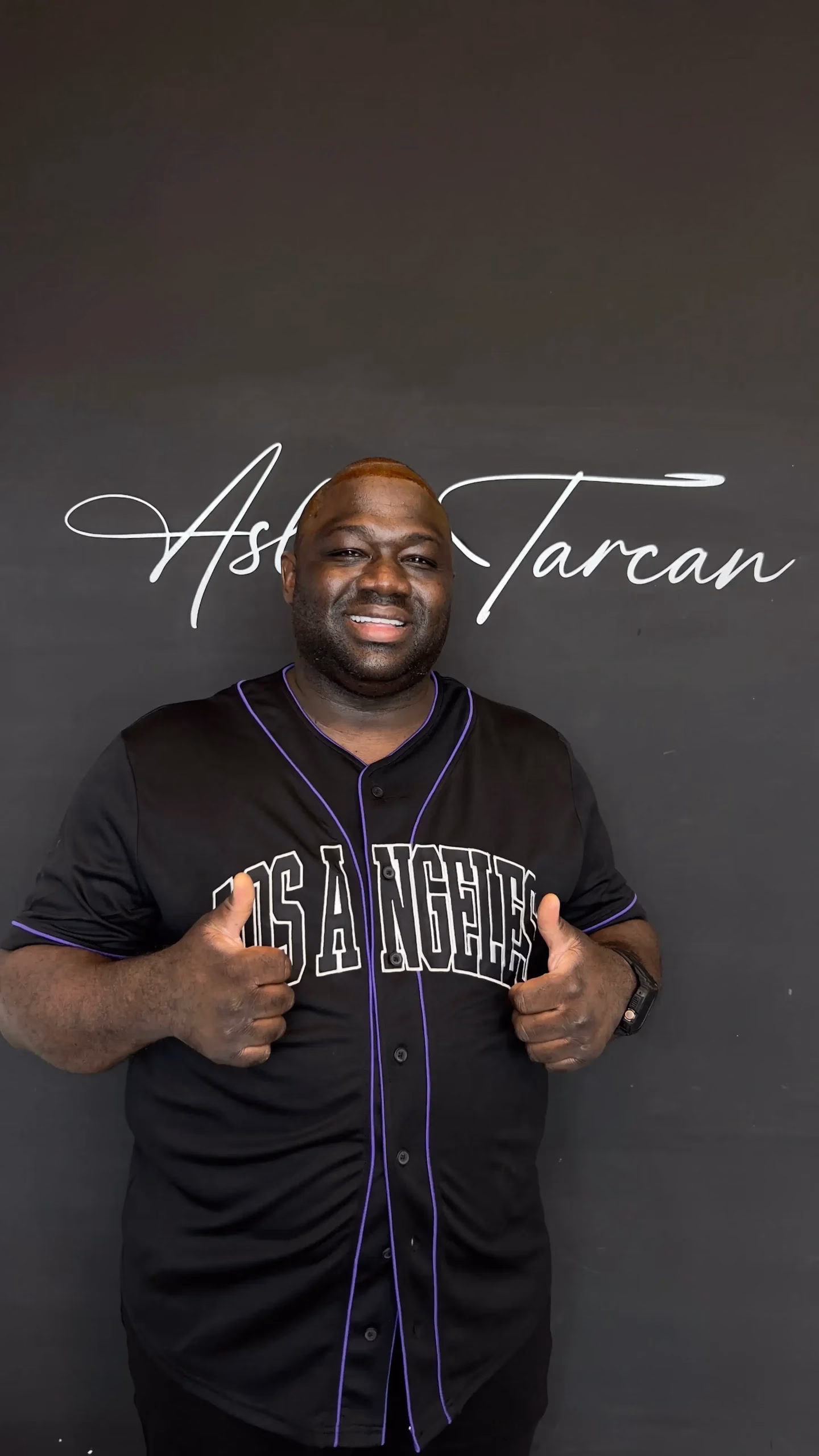Hombre sonriendo frente a una pared con el logo de una clínica de trasplante capilar, mostrando ambos pulgares hacia arriba. Luce una camiseta negra y parece satisfecho con el resultado del tratamiento.