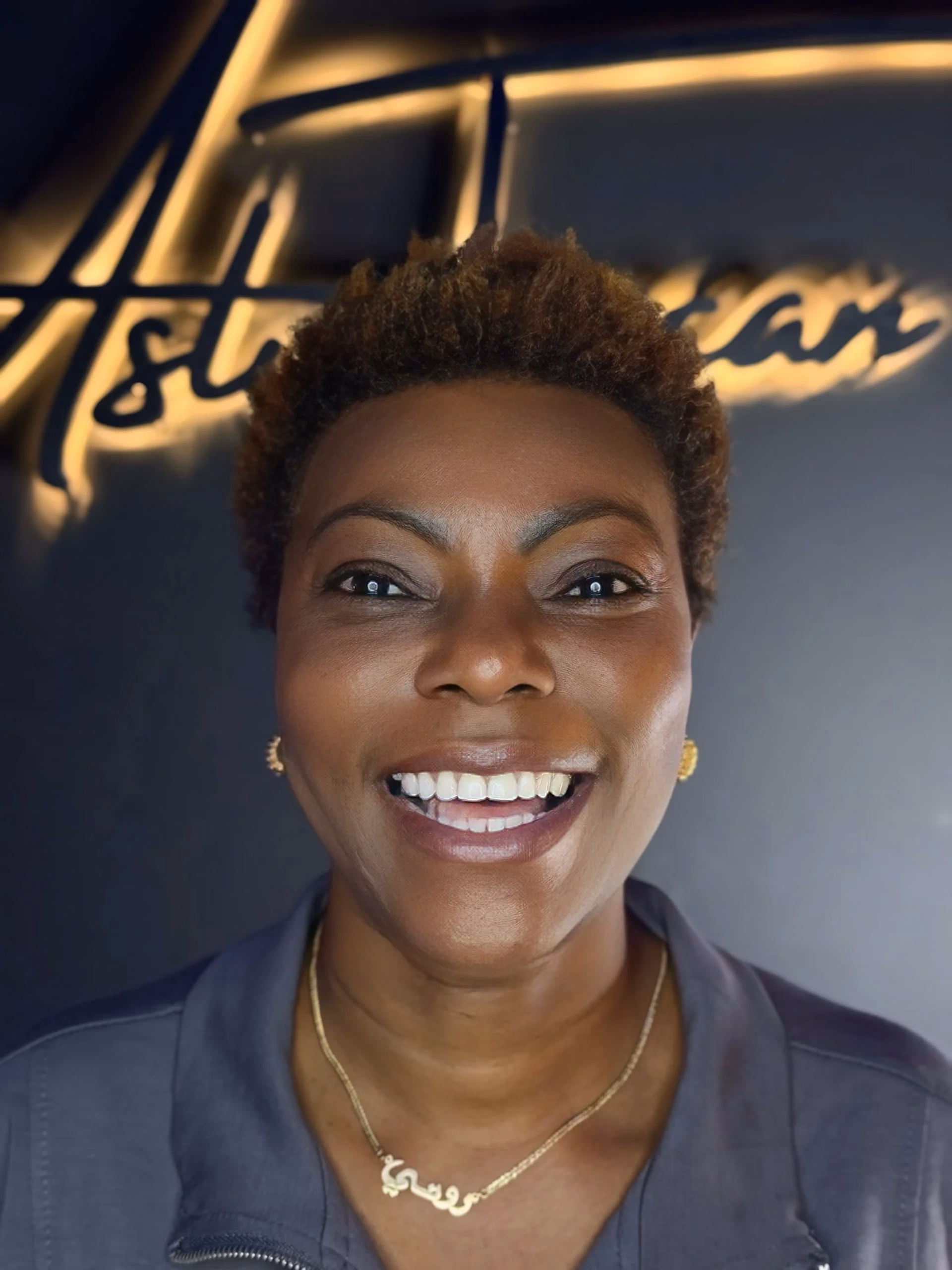 Una mujer sonriendo frente al logotipo iluminado de una clínica, con el cabello corto y rizado. Lleva pendientes dorados y un collar fino, y viste uniforme clínico gris, en un entorno profesional de estética capilar.