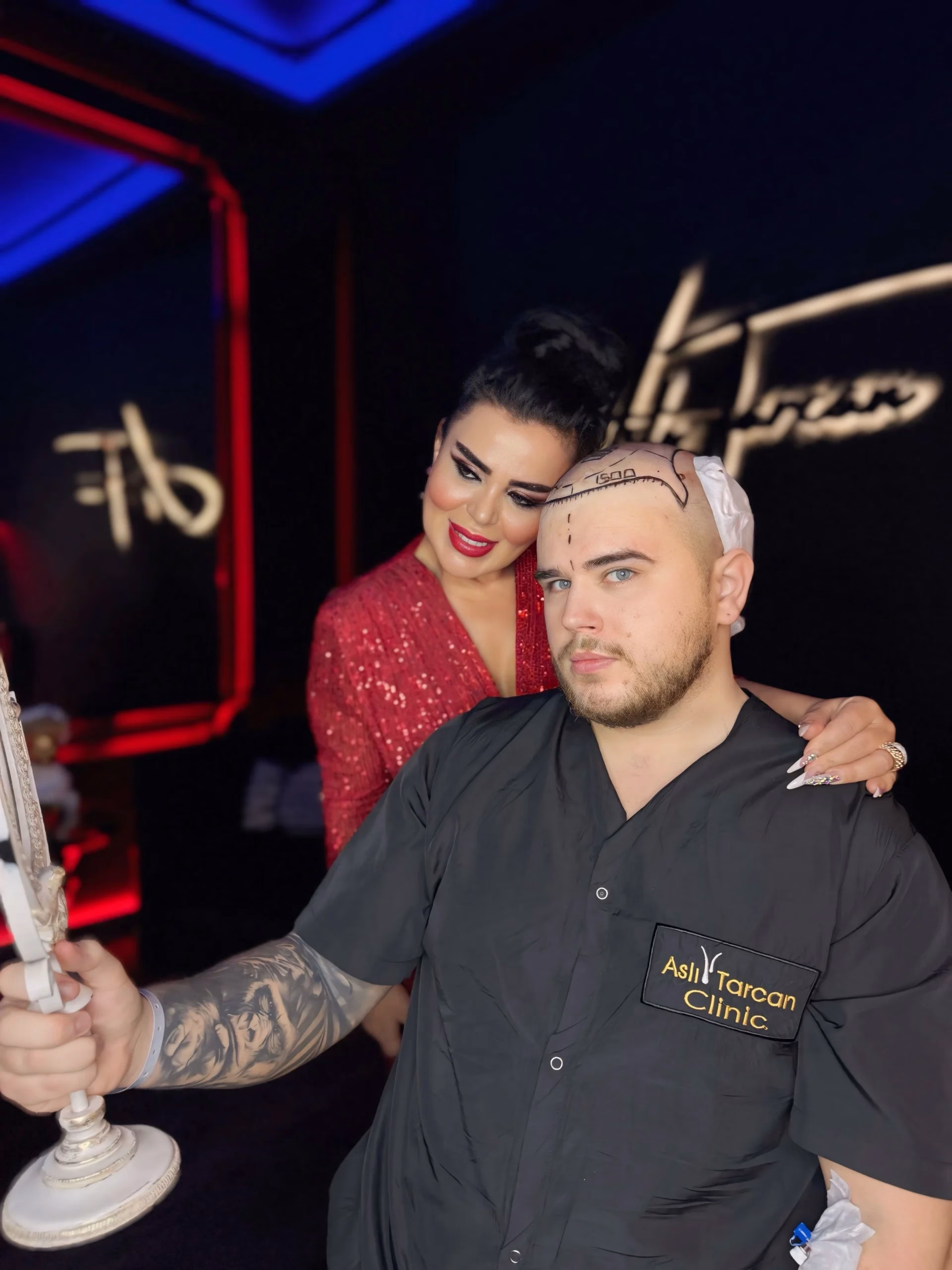 Un hombre en una clínica capilar observa su reflejo en un espejo de mano después de un procedimiento de trasplante de cabello; tiene marcas quirúrgicas visibles en la cabeza y vendaje. Viste una bata negra con el logo de la clínica. Una mujer con vestido rojo está detrás de él, sonriendo y apoyando la mano en su hombro en un ambiente elegante con luces tenues.