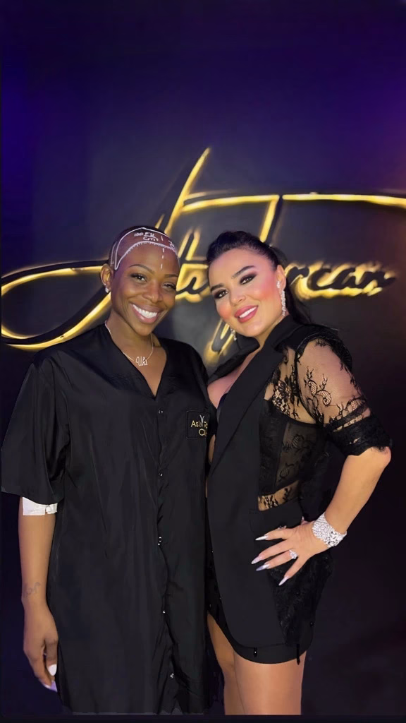 Dos mujeres posan sonrientes en una clínica estética frente a un fondo elegante con logotipo iluminado; una lleva una bata clínica negra y la otra un vestido negro con detalles de encaje.