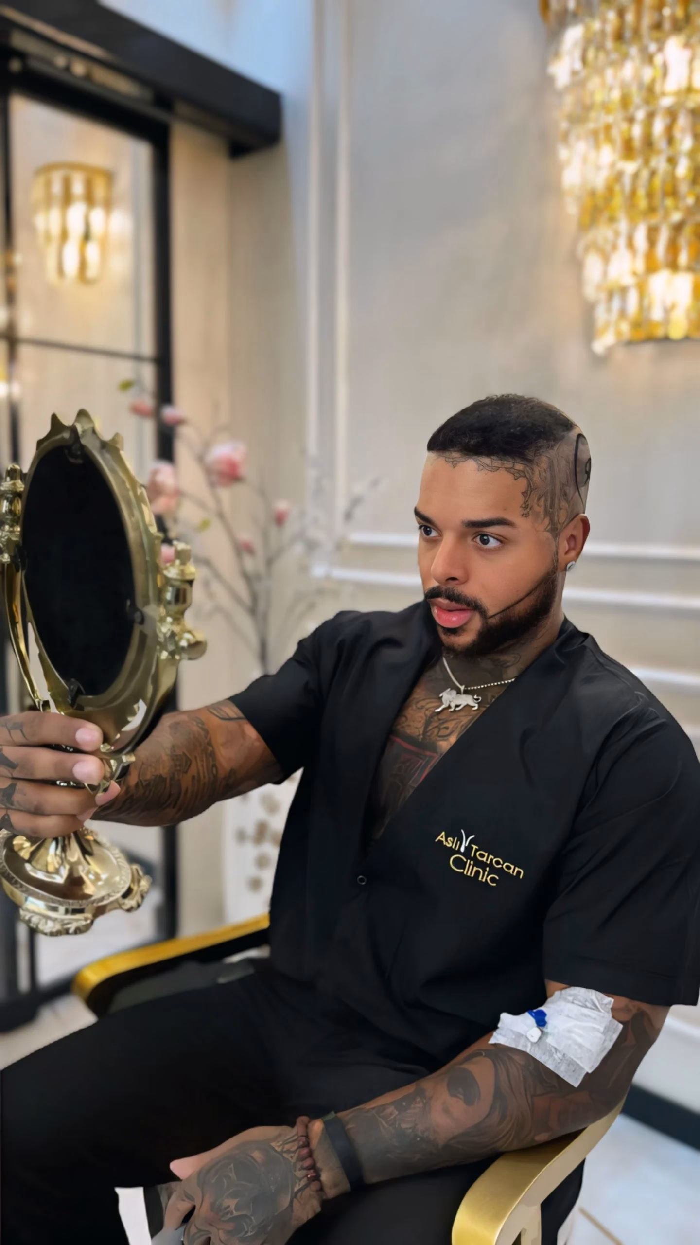 Un hombre joven con tatuajes observa su nuevo cabello en un espejo de mano dentro de una clínica elegante. Está sentado en una silla clínica, lleva una camisa negra con el logo de una clínica capilar y tiene un apósito médico en el brazo, lo que sugiere que acaba de someterse a un procedimiento de trasplante capilar.