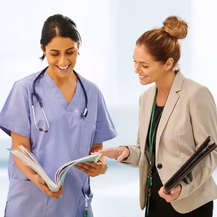 Profesional de la salud con bata médica y estetoscopio conversando sonriente con una mujer de negocios mientras revisan documentos en un entorno clínico luminoso.